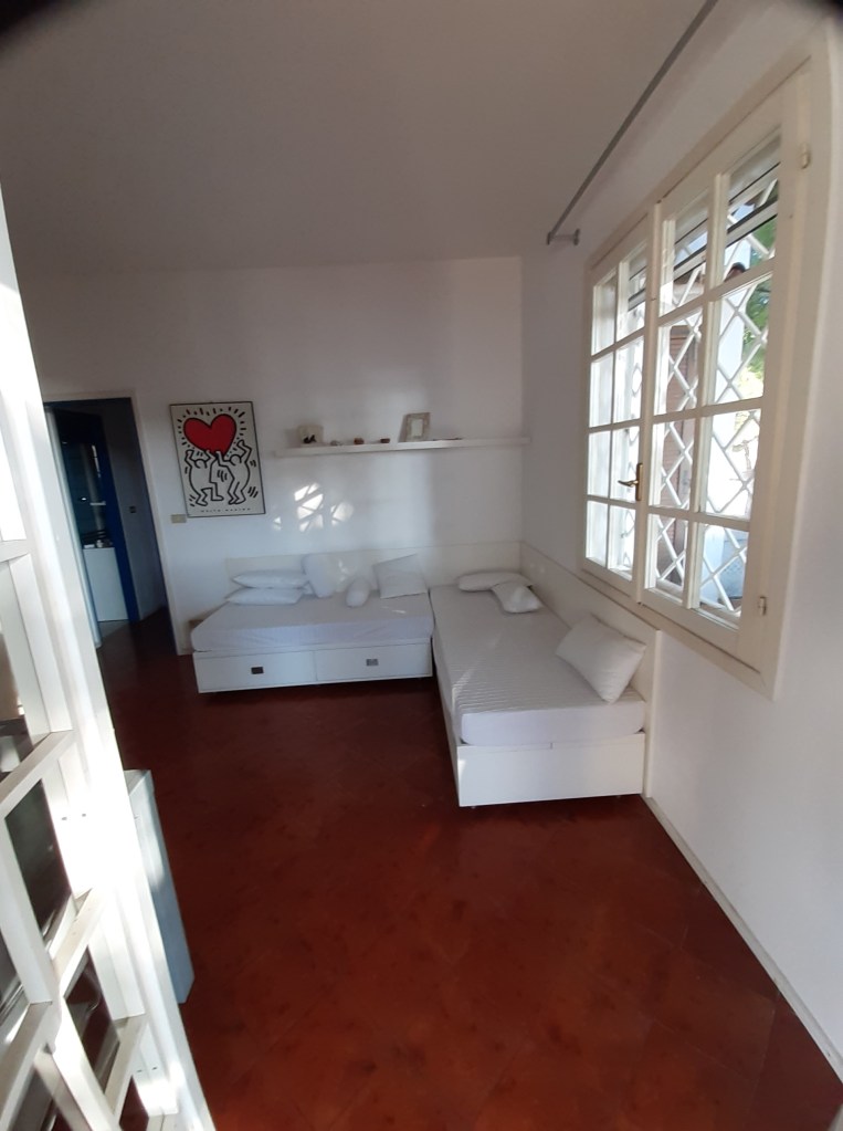 Ein gemütlicher Raum in einer Villa mit zwei Einzelbetten, weißen Wänden und einem großen Fenster mit Blick ins Freie. Kunstwerk an der Wand und heller Holzfußboden.