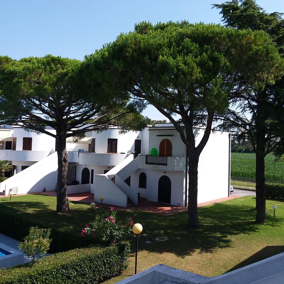 Exterior view of a charming white villa surrounded by trees and greenery, featuring steps leading to balconies and an inviting garden area.