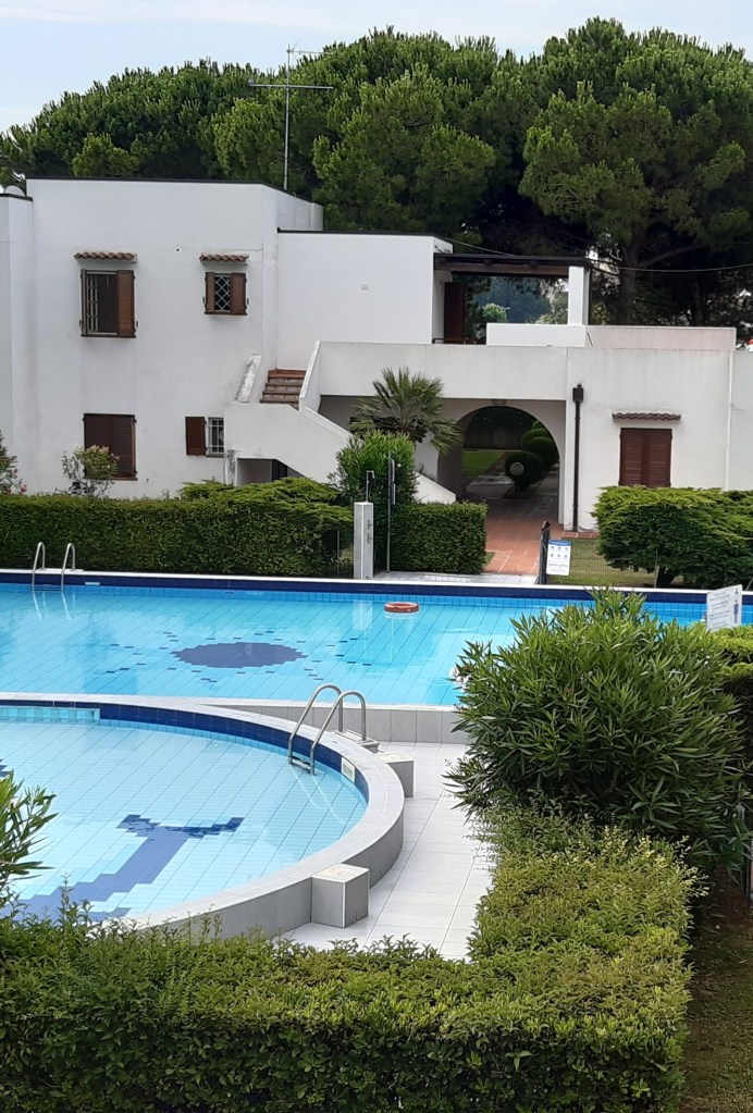 Vista dell'area piscine del residence, con una piscina circolare e un'altra rettangolare, circondate da verde e alberi.