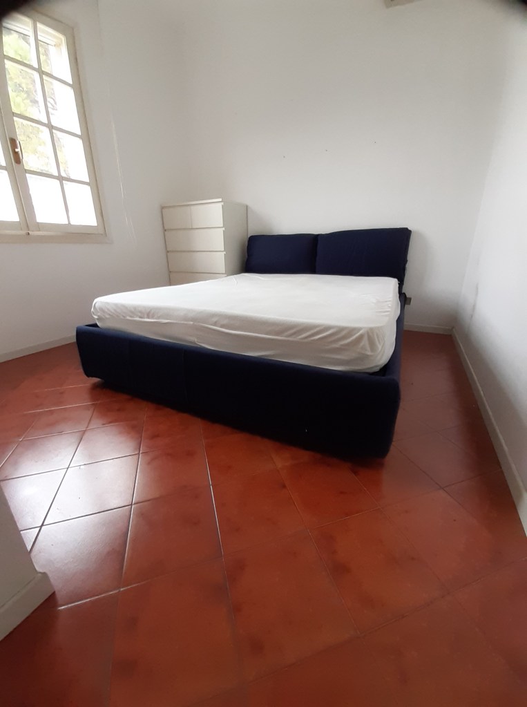 A bedroom showing a blue bed frame with a mattress, a white cabinet, and terracotta tiled flooring, illuminated by natural light from a window.