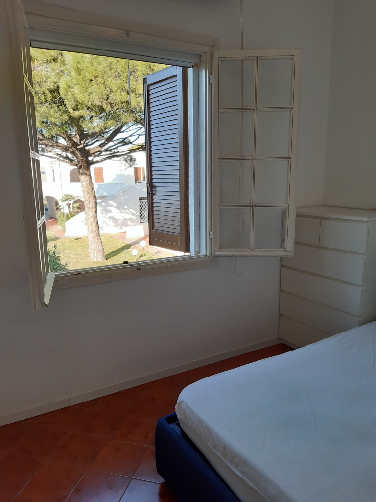 Interno di una camera da letto con un letto bianco e un armadio, vista da una finestra aperta che mostra un albero e il giardino del residence.