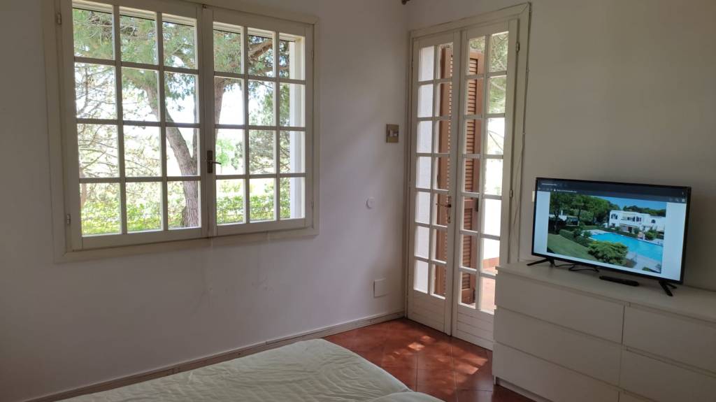 Interno di una camera da letto con finestra a griglia che mostra un albero all'esterno, TV su un mobile e porta a vetri che conduce all'esterno.