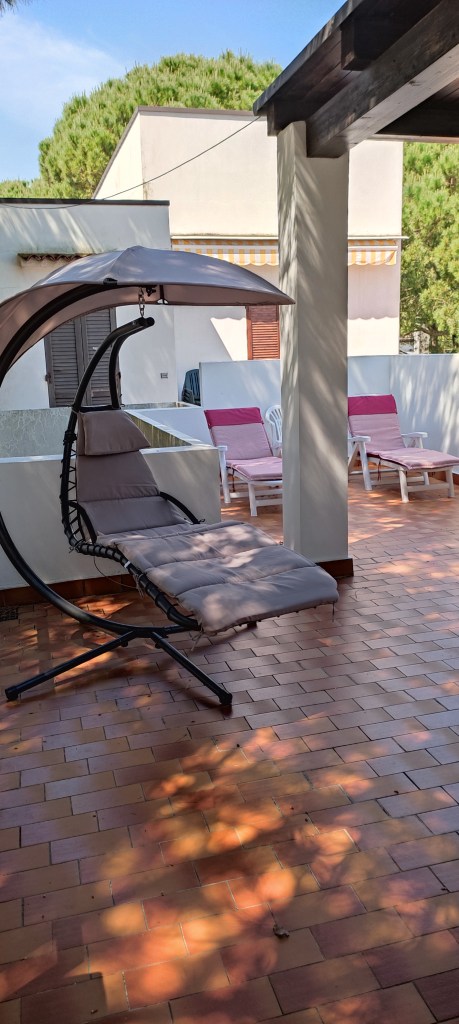 Terrazza di una villetta con un'amaca da esterno e sedie prendisole, circondata da alberi.