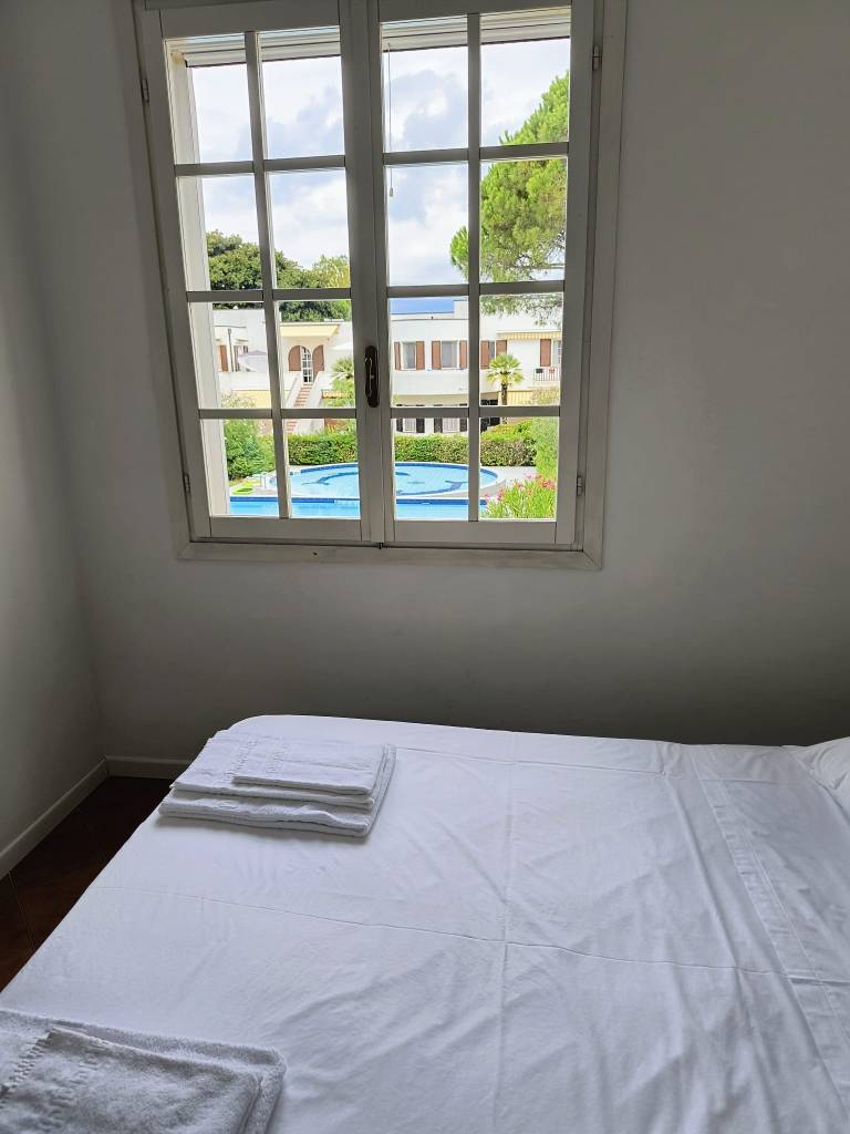 View from inside a villa room showing an open window with white curtains and a glimpse of a swimming pool and greenery outside.